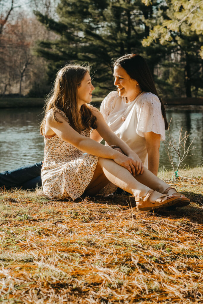 Mother and Daughter smile at each other in front of pond at horse barn during rustic family photos in the Philadelphia suburbs