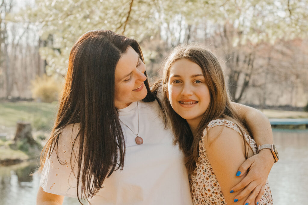 Mother looks on as daughter smiles at the camera at horse barn during barn family photos in the Philadelphia suburbs