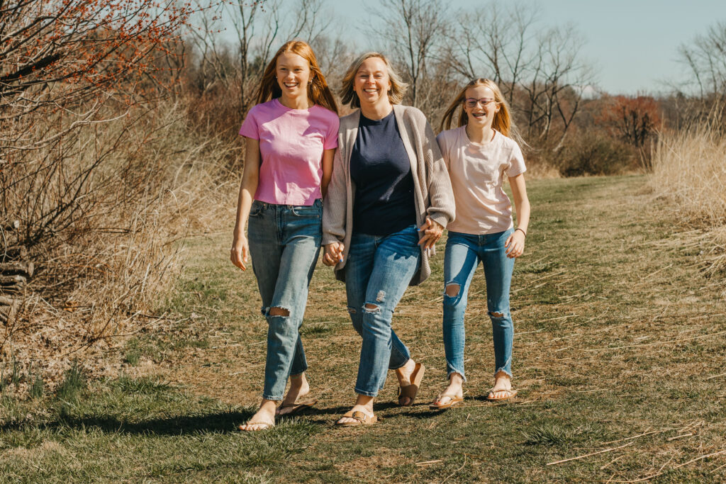 a mother and her two daughters walking down path, all laughing, during mother daughter photoshoot at marsh creek state park in the Philadelphia suburbs