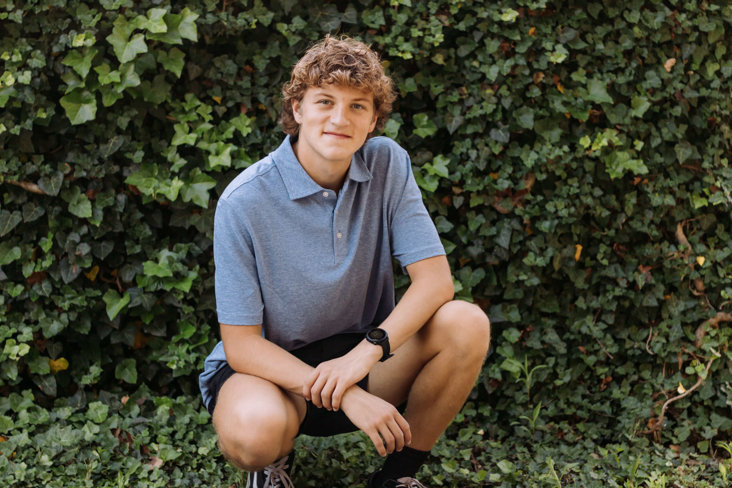 Male HS senior crouches in front of ivy wall during Senior Pictures in 2024